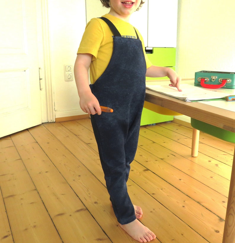 May include: A child wearing a blue denim overall with a yellow shirt and a brown crayon in their hand.