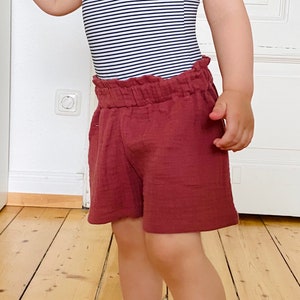 Op de afbeelding: Een kinderoutfit met een zwart-wit gestreepte badpak met ruches en een bijpassende strik. De outfit bevat bordeauxrode shorts. Geschikt voor warm weer en strandactiviteiten.