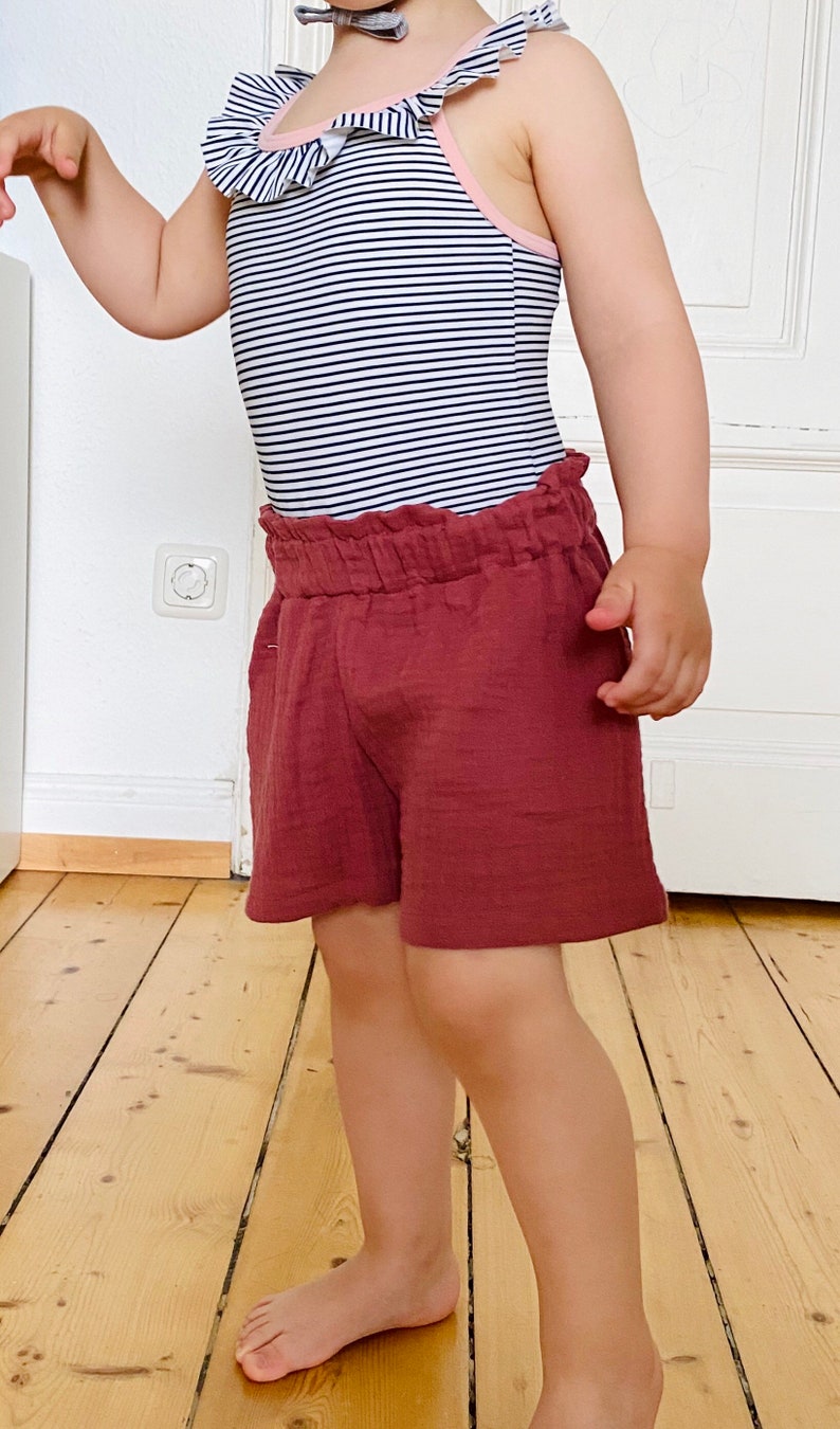 Op de afbeelding: Een kinderoutfit met een zwart-wit gestreepte badpak met ruches en een bijpassende strik. De outfit bevat bordeauxrode shorts. Geschikt voor warm weer en strandactiviteiten.