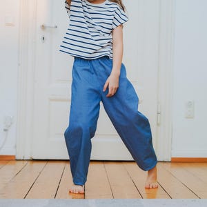 May include: A child wearing a striped short-sleeved shirt and blue pants. The shirt has horizontal navy blue stripes on a white background. The pants are a solid light blue color. The child is standing on a wooden floor.