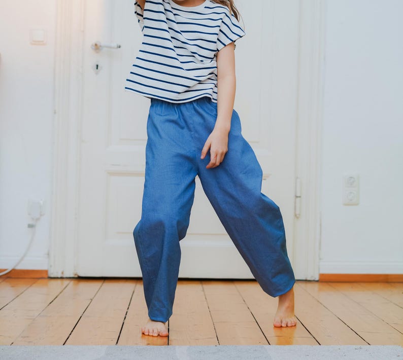 May include: A child wearing a striped short-sleeved shirt and blue pants. The shirt has horizontal navy blue stripes on a white background. The pants are a solid light blue color. The child is standing on a wooden floor.