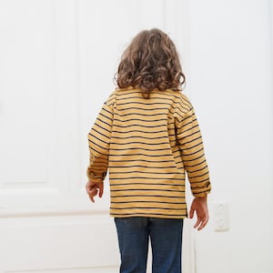 Puede incluir: Un ni&ntilde;o con una camisa de manga larga a rayas amarillas y azul marino y pantalones vaqueros azules.