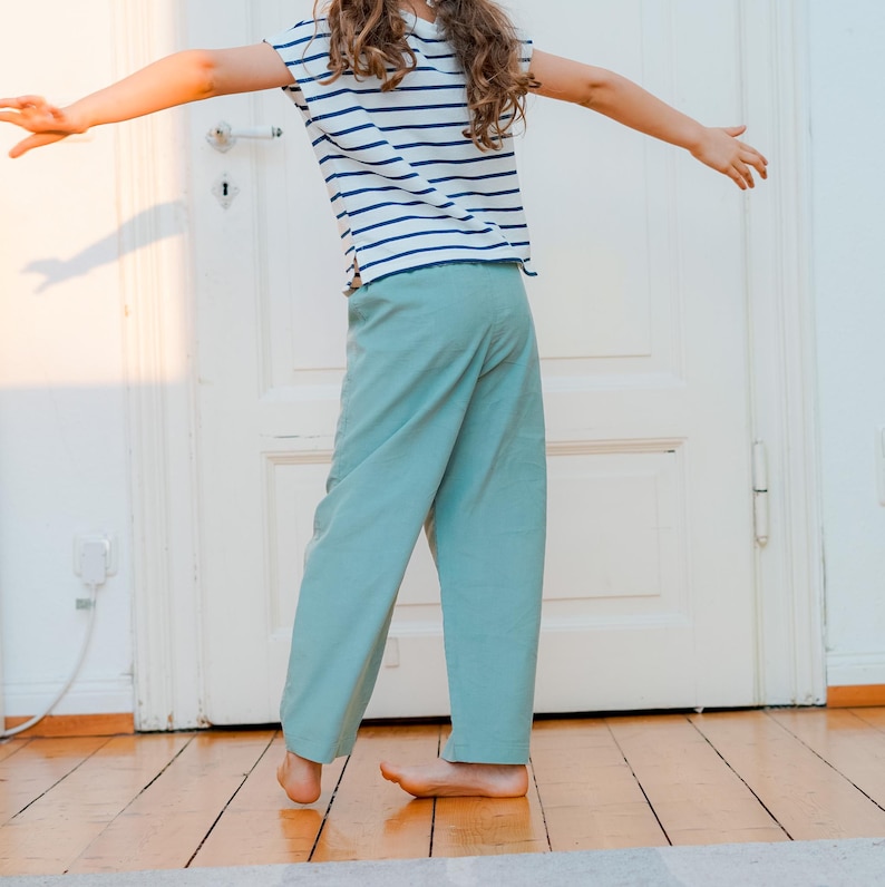 May include: A child wearing a striped t-shirt with blue and white stripes and light green pants. The child is standing on a wooden floor in front of a white door, arms outstretched.