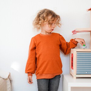 May include: A young person with curly blonde hair wearing a burnt orange long-sleeved shirt and blue jeans plays with a wooden toy house. The house has a blue and white striped awning and a gray door.