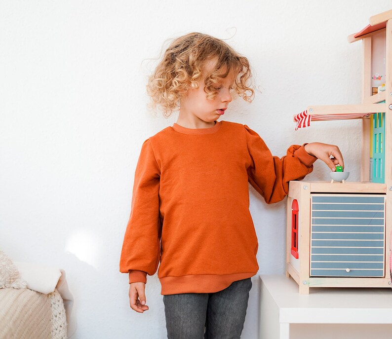 May include: A young person with curly blonde hair wearing a burnt orange long-sleeved shirt and blue jeans plays with a wooden toy house. The house has a blue and white striped awning and a gray door.