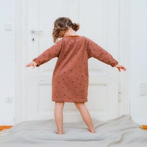 May include: A young person wearing a brown dress with a pattern of small squares. The dress has long sleeves and a loose fit. The person is standing on a light brown rug in front of a white door.