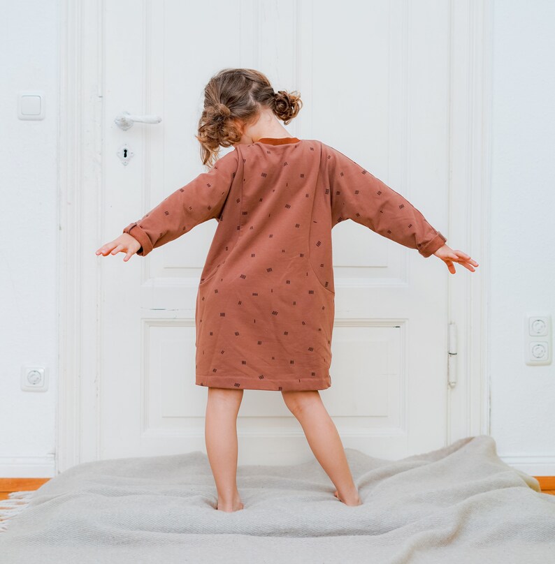 May include: A young person wearing a brown dress with a pattern of small squares. The dress has long sleeves and a loose fit. The person is standing on a light brown rug in front of a white door.