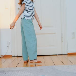 May include: A person wearing a striped, short-sleeved shirt and light blue pants. The person is standing on a wooden floor in front of a white door. The pants are a solid color, and the shirt has horizontal stripes.