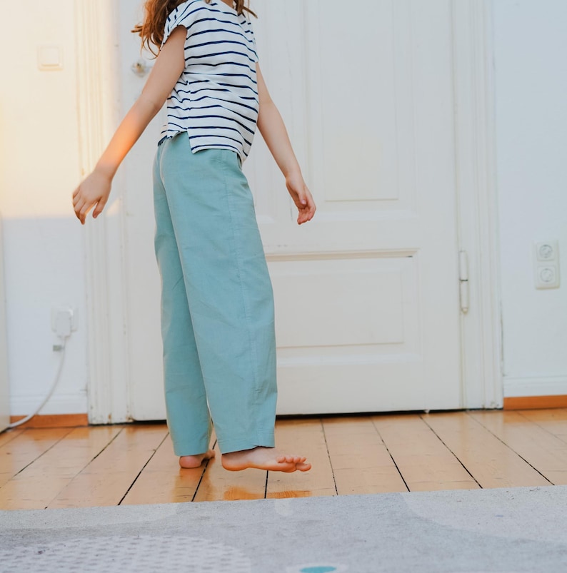 May include: A person wearing a striped, short-sleeved shirt and light blue pants. The person is standing on a wooden floor in front of a white door. The pants are a solid color, and the shirt has horizontal stripes.