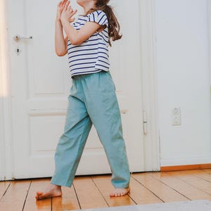 May include: A child wearing a striped short-sleeve shirt and light blue pants. The shirt has horizontal navy blue stripes on a white background. The pants are a solid light blue color. The child is standing on a wooden floor.