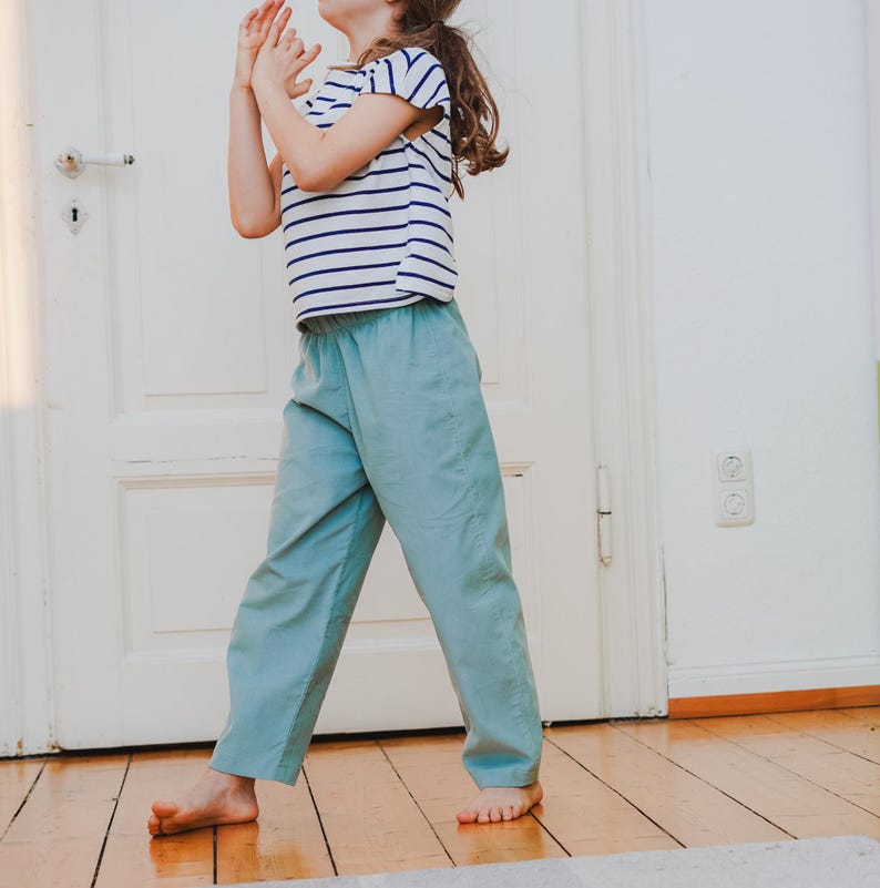 May include: A child wearing a striped short-sleeve shirt and light blue pants. The shirt has horizontal navy blue stripes on a white background. The pants are a solid light blue color. The child is standing on a wooden floor.