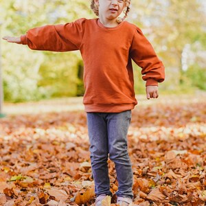 May include: A young person wearing a rust-colored sweatshirt with puffed sleeves and blue jeans. The person is standing in a pile of fallen autumn leaves.