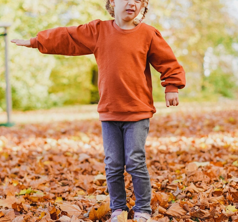 May include: A young person wearing a rust-colored sweatshirt with puffed sleeves and blue jeans. The person is standing in a pile of fallen autumn leaves.