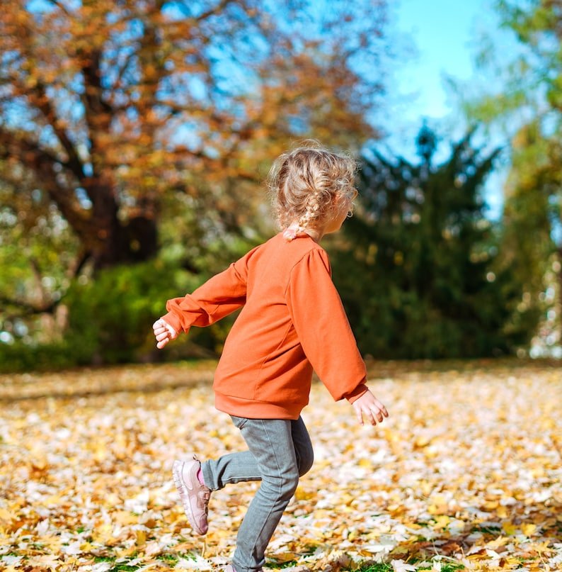 May include: A young girl with blonde hair in a braided ponytail wearing a bright orange long-sleeved shirt and grey jeans runs through a field of fallen autumn leaves.