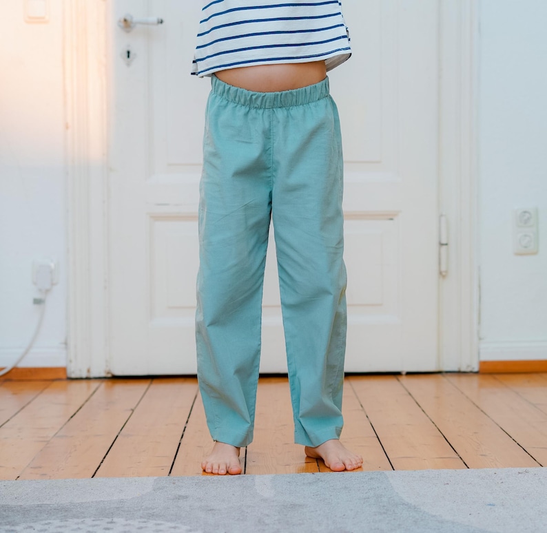 May include: A child wearing light blue pants with an elastic waistband and a striped shirt. The pants are a solid color and appear to be made of a lightweight fabric. The child is standing on a wooden floor.