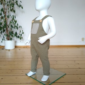 May include: A green and brown baby overall with a patch pocket and wooden buttons. The overall is on a white mannequin.