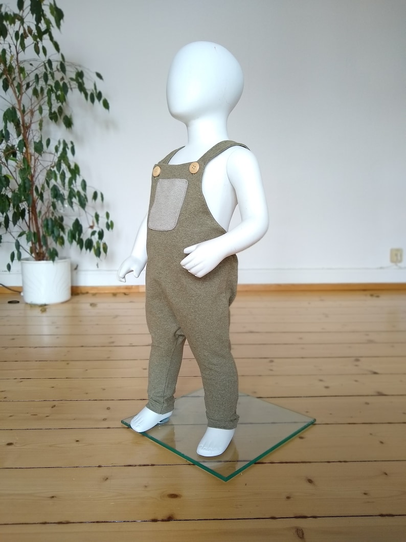 May include: A green and brown baby overall with a patch pocket and wooden buttons. The overall is on a white mannequin.