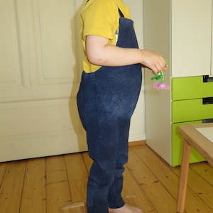 May include: A young child wearing a yellow short-sleeved shirt and blue denim overalls. The child is standing on a wooden platform and holding a pink and green plastic flower.