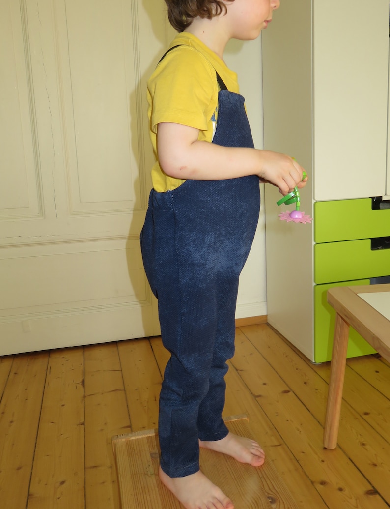 May include: A young child wearing a yellow short-sleeved shirt and blue denim overalls. The child is standing on a wooden platform and holding a pink and green plastic flower.