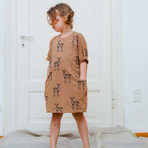 May include: A brown dress with a deer print pattern. The dress has short sleeves and two pockets. The dress is worn by a young person standing on a light brown rug.