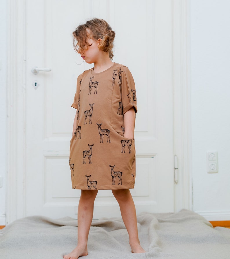 May include: A brown dress with a deer print pattern. The dress has short sleeves and two pockets. The dress is worn by a young person standing on a light brown rug.