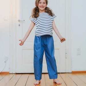 May include: A young person wearing a white and navy blue striped t-shirt and blue pants. The t-shirt has short sleeves and a round neckline. The pants are a solid blue color and have a straight leg. The person is standing on a wooden floor.