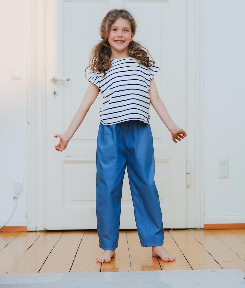 May include: A young person wearing a white and navy blue striped t-shirt and blue pants. The t-shirt has short sleeves and a round neckline. The pants are a solid blue color and have a straight leg. The person is standing on a wooden floor.