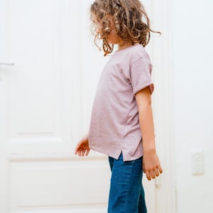Puede incluir: Una persona joven con una camiseta rosa claro con una textura sutil y pantalones vaqueros azules. La persona est&aacute; de pie frente a una puerta blanca.