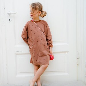 May include: A young girl wearing a brown dress with a white pattern and a pink purse. The dress has a pocket on the left side. The girl is standing in front of a white door.