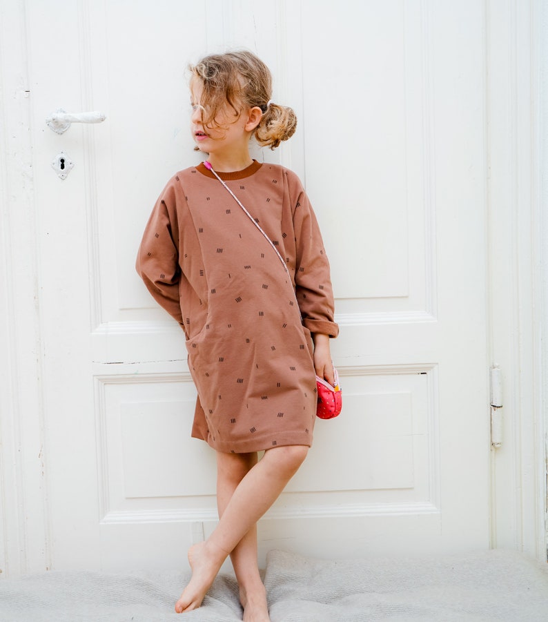 May include: A young girl wearing a brown dress with a white pattern and a pink purse. The dress has a pocket on the left side. The girl is standing in front of a white door.