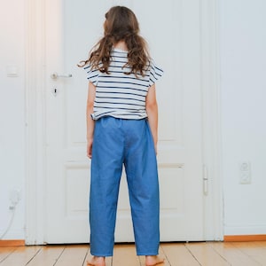 May include: A child wearing a striped white and navy blue t-shirt and blue pants. The child is standing in front of a white door on a wooden floor. The t-shirt has short sleeves and the pants are loose-fitting.