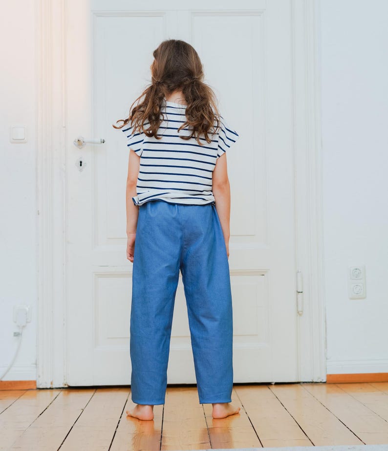May include: A child wearing a striped white and navy blue t-shirt and blue pants. The child is standing in front of a white door on a wooden floor. The t-shirt has short sleeves and the pants are loose-fitting.