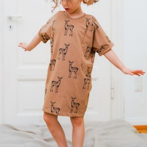 May include: A young person wearing a brown short-sleeved dress with a black deer print. The dress has a pocket on the left side.