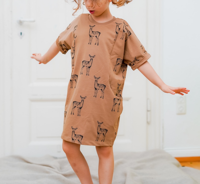 May include: A young person wearing a brown short-sleeved dress with a black deer print. The dress has a pocket on the left side.