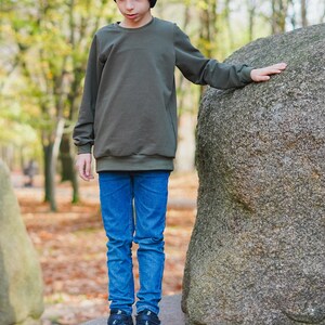 May include: A young person wearing a dark green sweatshirt and blue jeans stands in front of a large, gray rock. The sweatshirt has a crew neck and long sleeves.