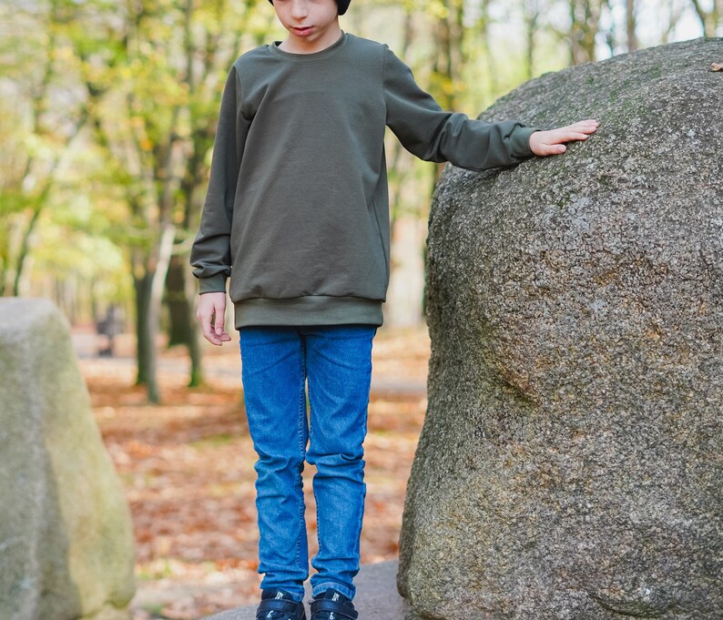 May include: A young person wearing a dark green sweatshirt and blue jeans stands in front of a large, gray rock. The sweatshirt has a crew neck and long sleeves.