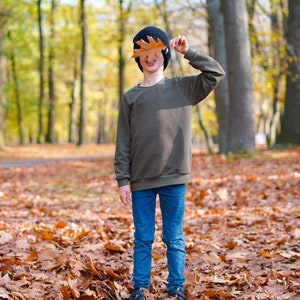 May include: A young person wearing a dark green sweater, blue jeans, and a black beanie holds a brown leaf in front of their face. They are standing in a forest with fallen leaves on the ground.