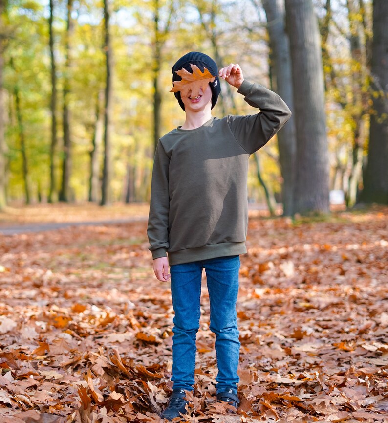 May include: A young person wearing a dark green sweater, blue jeans, and a black beanie holds a brown leaf in front of their face. They are standing in a forest with fallen leaves on the ground.