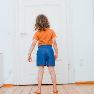 Op de afbeelding: Een kind draagt een oranje t-shirt met witte randen en een blauwe korte broek. Het kind staat voor een witte deur op een houten vloer. Het t-shirt heeft korte mouwen en een ronde halslijn.