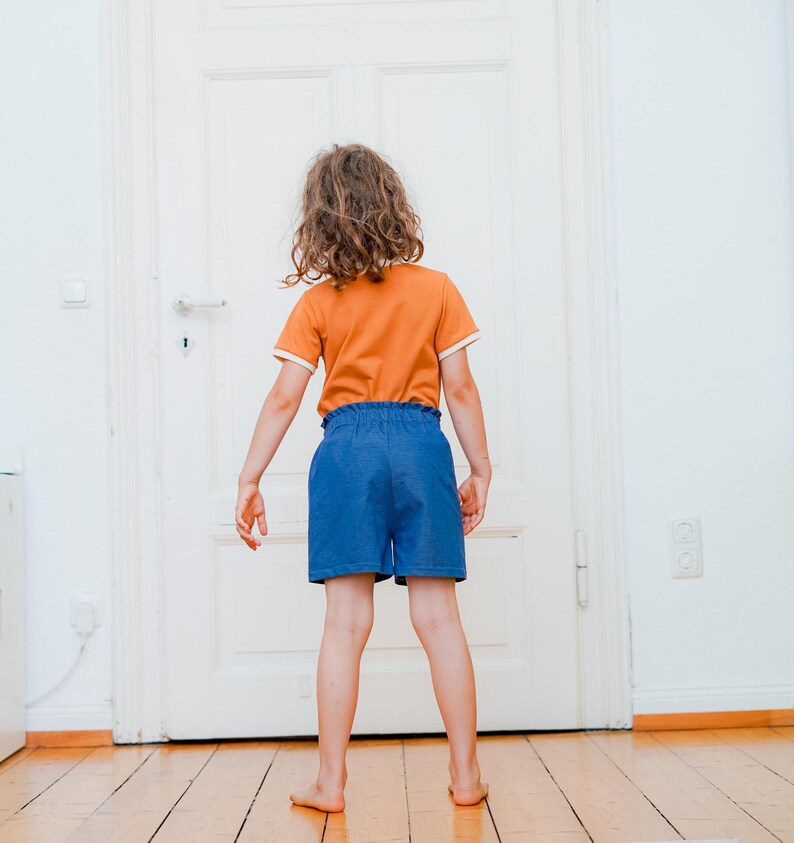 Op de afbeelding: Een kind draagt een oranje t-shirt met witte randen en een blauwe korte broek. Het kind staat voor een witte deur op een houten vloer. Het t-shirt heeft korte mouwen en een ronde halslijn.