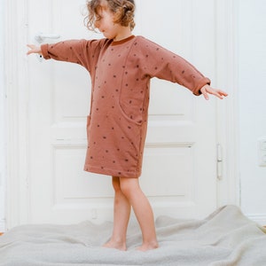 May include: A young person wearing a brown dress with a white and brown pattern. The dress has a large pocket on the front. The person is standing on a light brown rug.