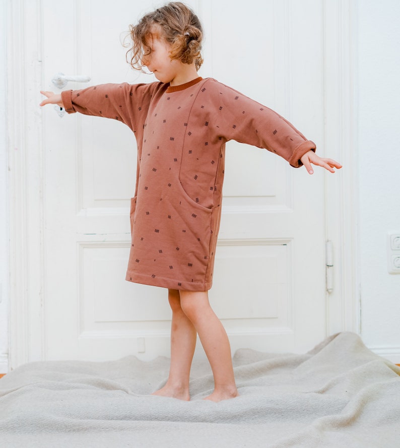 May include: A young person wearing a brown dress with a white and brown pattern. The dress has a large pocket on the front. The person is standing on a light brown rug.