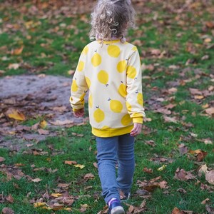May include: A young person wearing a yellow and white sweater with a lemon print. The sweater has long sleeves and a crew neck. The person is wearing blue jeans and is standing in a grassy area with fallen leaves.