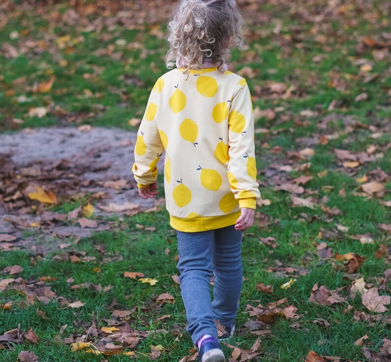 May include: A young person wearing a yellow and white sweater with a lemon print. The sweater has long sleeves and a crew neck. The person is wearing blue jeans and is standing in a grassy area with fallen leaves.