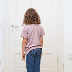 Puede incluir: Una persona joven con una camiseta rosa moteada y pantalones vaqueros azules se encuentra frente a una puerta blanca.