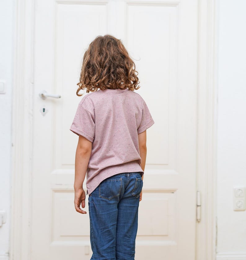 Puede incluir: Una persona joven con una camiseta rosa moteada y pantalones vaqueros azules se encuentra frente a una puerta blanca.