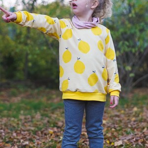May include: A young person wearing a yellow sweatshirt with a lemon print and blue jeans. The sweatshirt has a yellow band around the bottom.