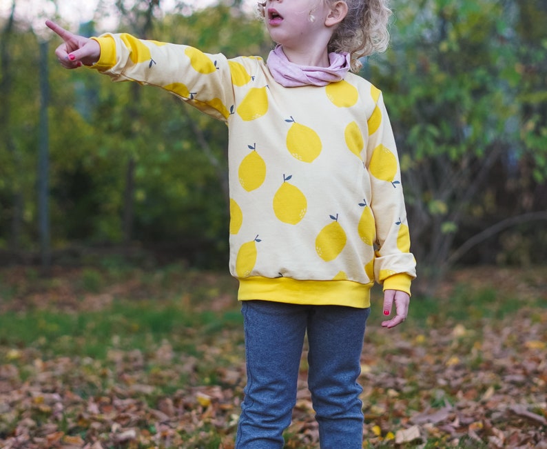 May include: A young person wearing a yellow sweatshirt with a lemon print and blue jeans. The sweatshirt has a yellow band around the bottom.