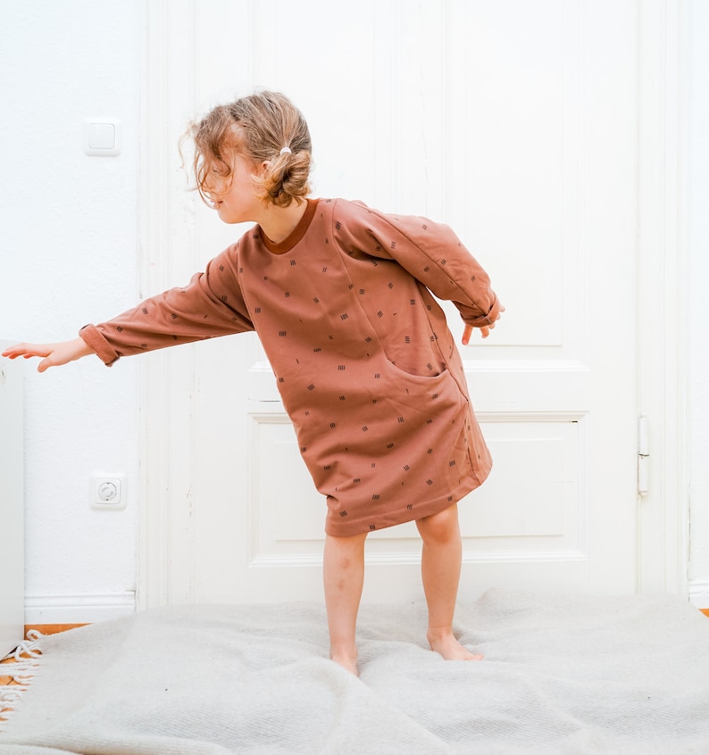 May include: A young person wearing a brown long-sleeved dress with a small pattern of brown squares. The dress has a large pocket on the left side. The person is standing on a light brown rug.
