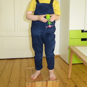 May include: A child wearing a blue denim overall jumpsuit with yellow straps over a yellow short-sleeved shirt. The child is standing on a wooden box.
